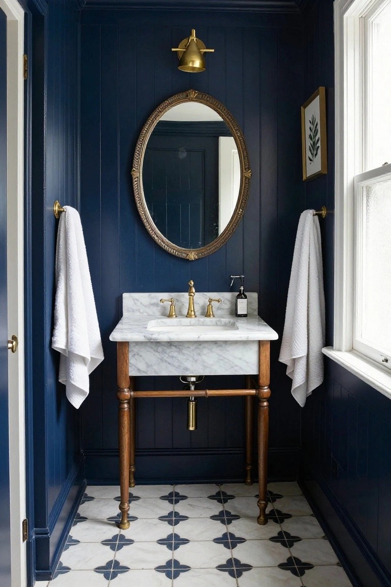 Small powder room with navy blue vertical paneled walls, wooden pedestal sink topped in white marble, oval gold-framed mirror, gold wall sconce, white towels on brass towel bars, soap dispenser, and white floor tiles patterned in navy quatrefoils.