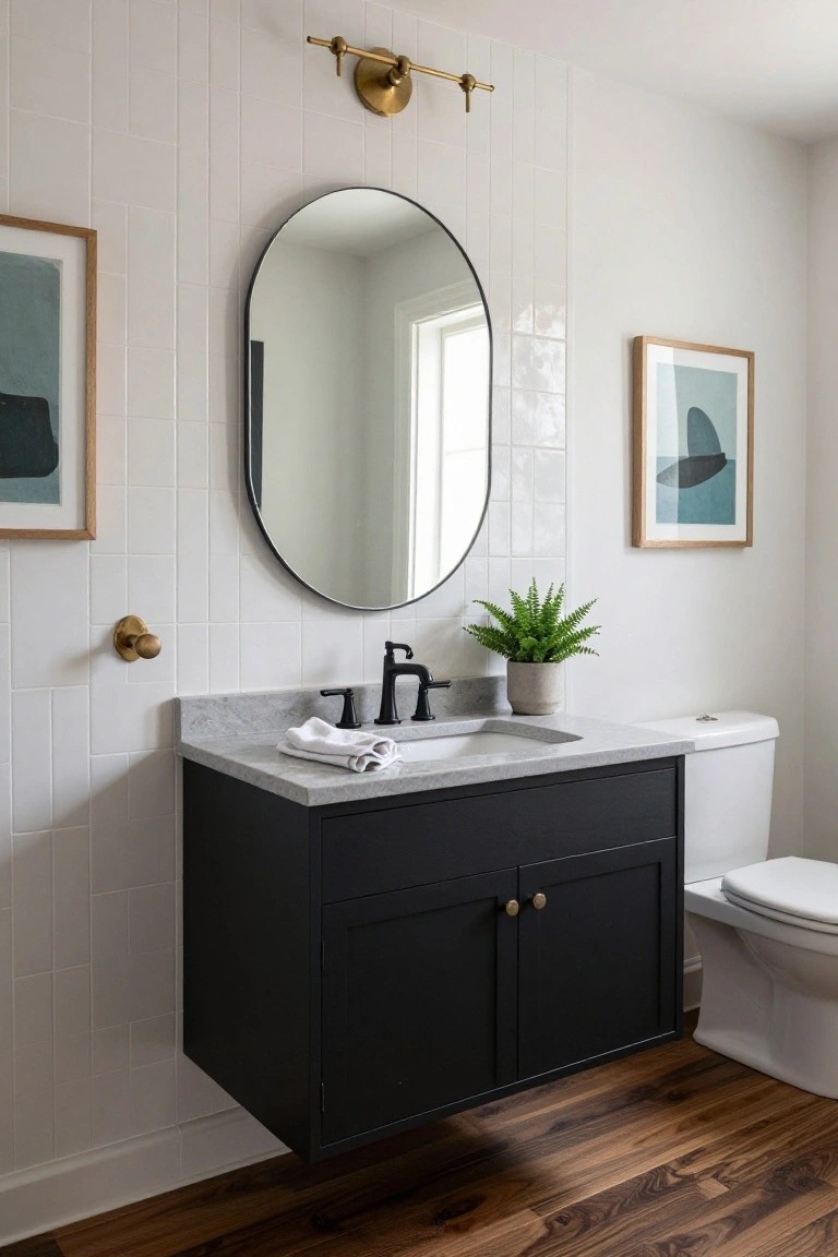 Modern bathroom with white subway tile walls, black floating vanity, gray quartz countertop, matte black faucet, oval black-framed mirror, brass sconce and knobs, potted fern, abstract framed art, white toilet, and wood plank flooring.