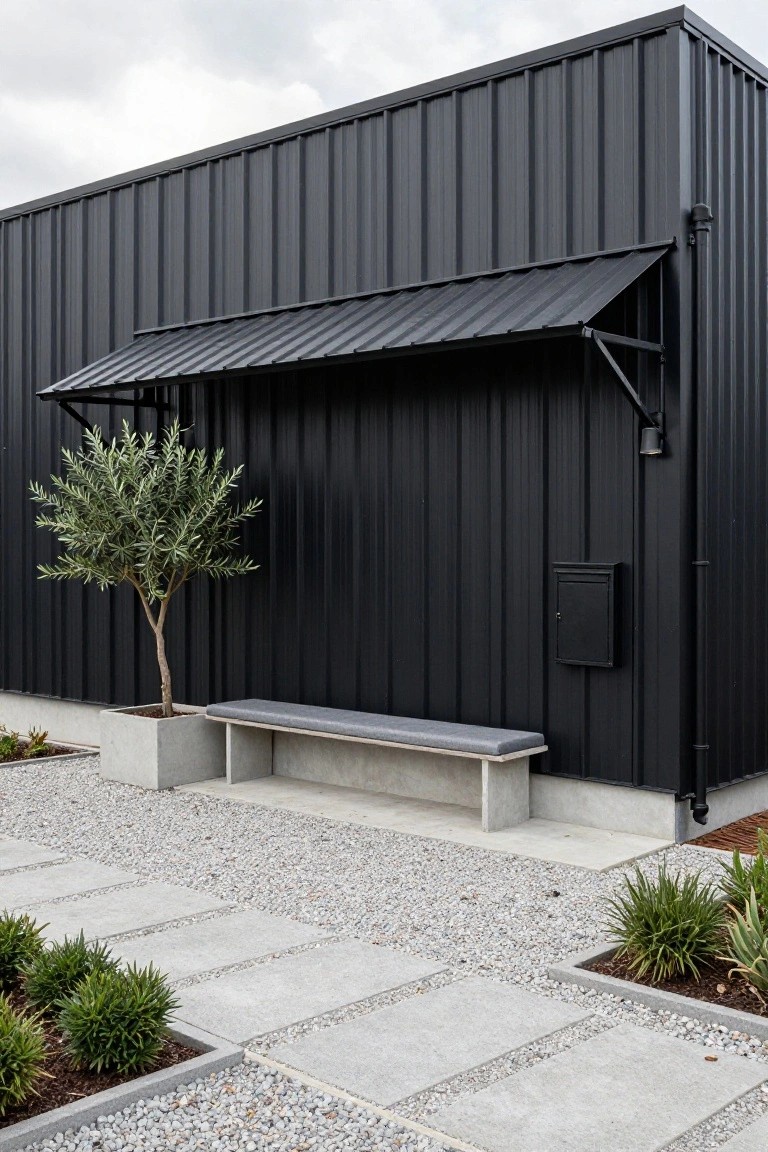 Black corrugated metal barn-style house exterior with awning, concrete bench, potted olive tree, black mailbox, gravel path with concrete pavers, and low plants.
