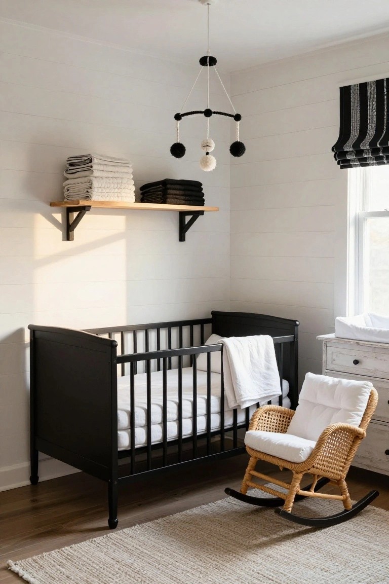 A black metal crib with white bedding stands in a white shiplap-walled nursery next to a white changing dresser, rattan rocking chair with white cushion, black floating shelves with stacked towels, black and white roman shade on the window, seagrass rug on wood floor.