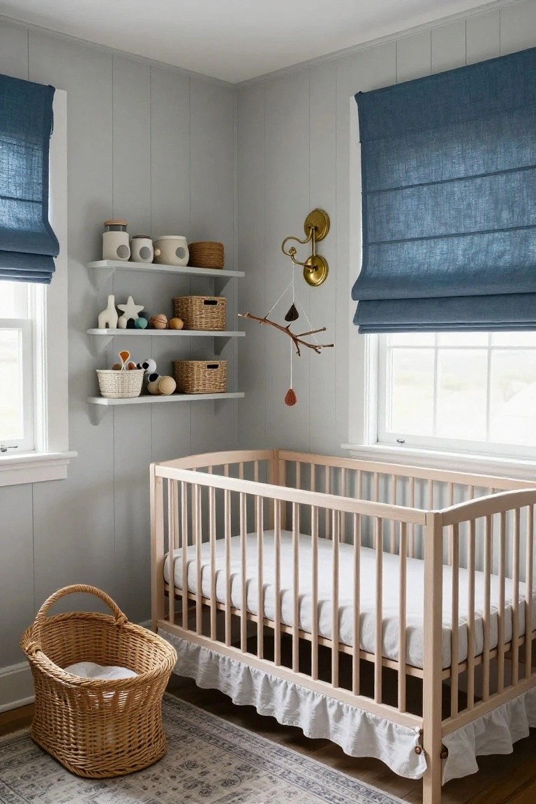 Corner of a light gray shiplap nursery with a natural wood crib having white bedding and ruffled skirt, a wicker bassinet on the floor nearby, floating white shelves holding toys plants and baskets, blue linen roman shades on two windows, and a patterned rug.