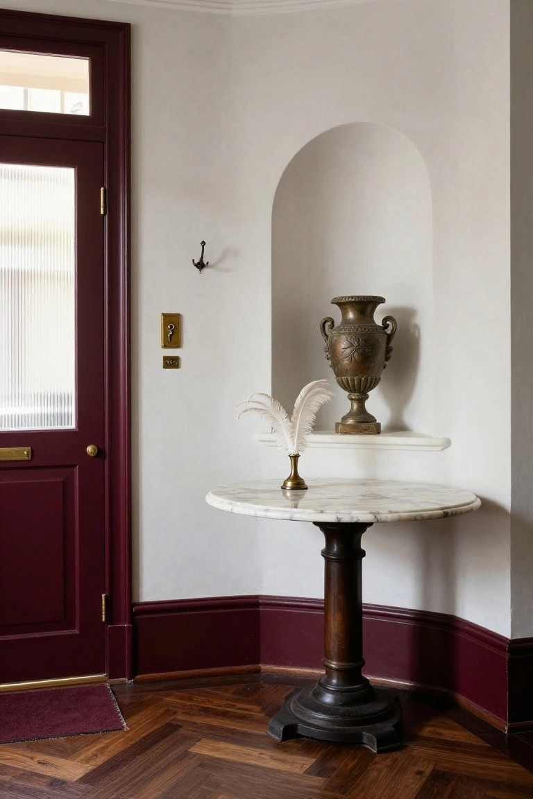 Foyer corner with burgundy paneled front door, white walls including an arched niche with brass urn and white feather on a shelf, small round white marble-topped pedestal table with brass candlestick, brass wall light, and herringbone parquet wood floor.