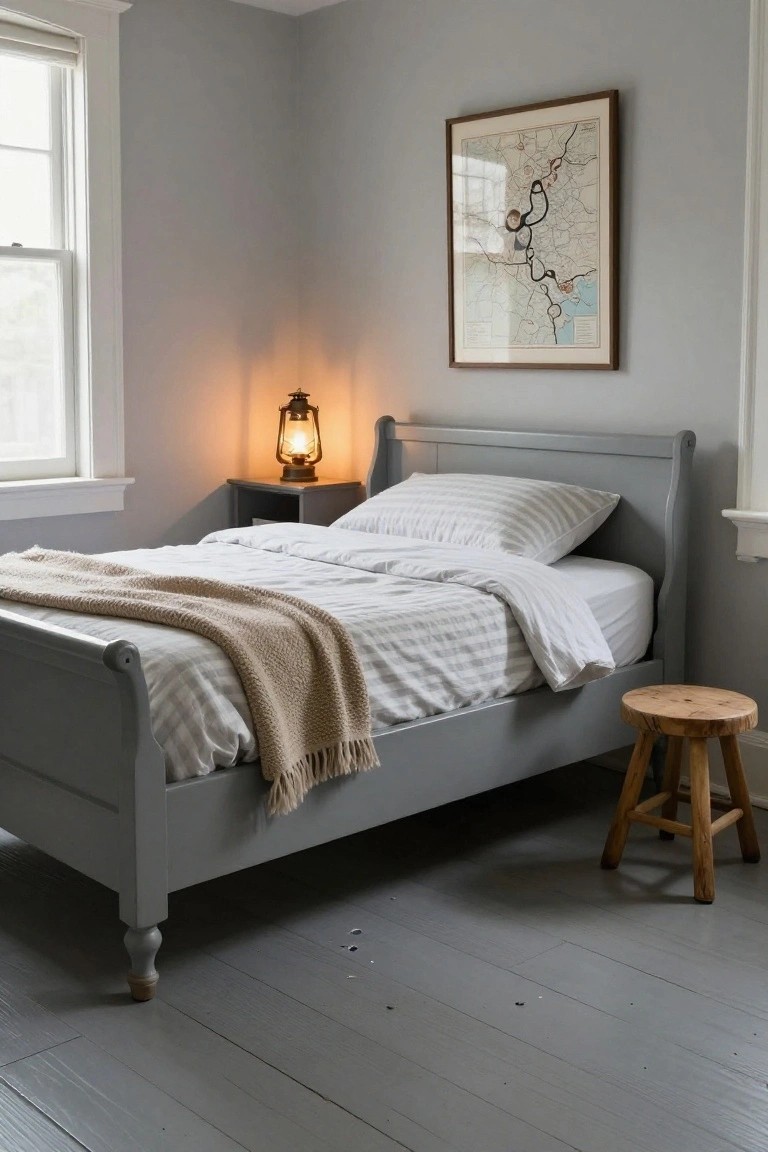 A gray bedroom with painted gray sleigh bed draped in white striped sheets and tan throw blanket, wooden stool nearby, lantern lamp on small table, framed map on wall, double-hung window, and wide plank gray painted wood floors.