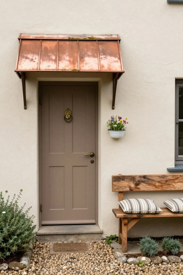 Gray paneled front door with brass leaf-shaped knocker and handle under a weathered copper awning on a white stucco wall, rustic wooden bench with striped cushions beside it, hanging white pot of flowers, gravel path, and low shrubs.
