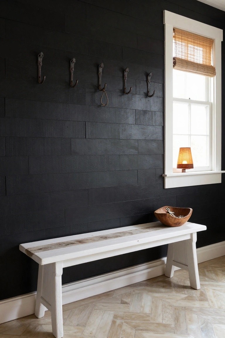 Entryway with black shiplap wall holding five iron hooks, white wooden bench next to it with a wooden bowl containing keys, window with bamboo shade and small lamp on sill, and light herringbone flooring.