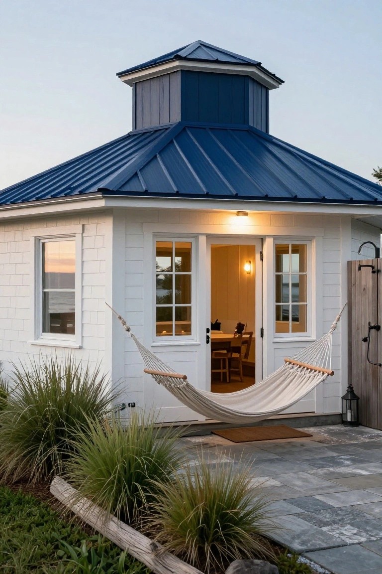 Small white shingle-sided barn-style building with blue metal roof and dark blue cupola, open double doors revealing wooden interior with table and chairs, white hammock hanging on porch, stone patio, grasses, driftwood, outdoor shower, and ocean view at dusk.
