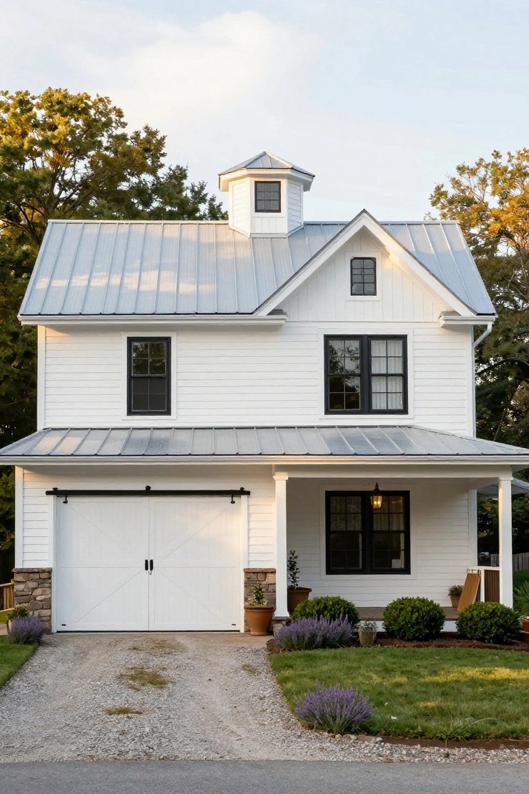 White two-story barn-style house with gray standing seam metal roof, octagonal cupola, black-framed windows, double garage doors, covered front porch, gravel driveway, and lavender plantings in the yard.