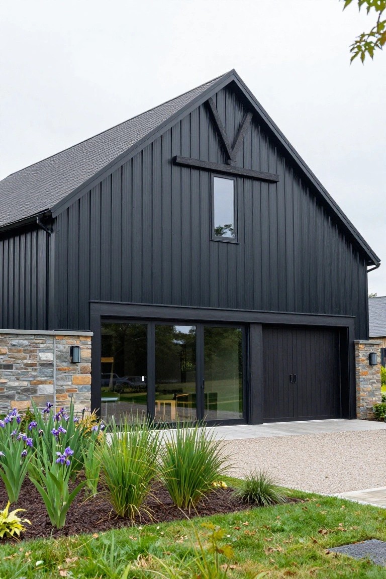 Black vertical board-and-batten sided barn-style house exterior with gabled roof, large glass-paneled garage door, stone accent piers, and front yard landscaping with purple irises, grasses, and gravel driveway.