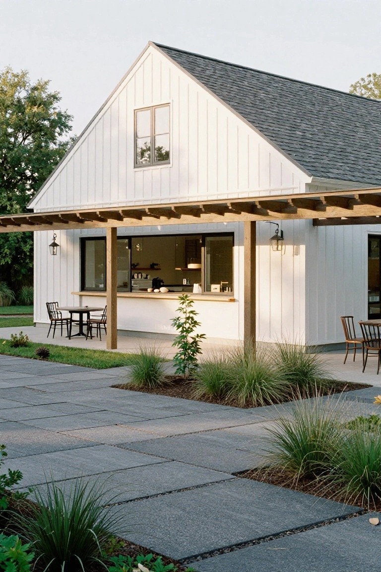 White shiplap barn-style house with wooden pergola-covered porch, kitchen pass-through counter, small round table with chairs, concrete paver patio, and ornamental grasses.
