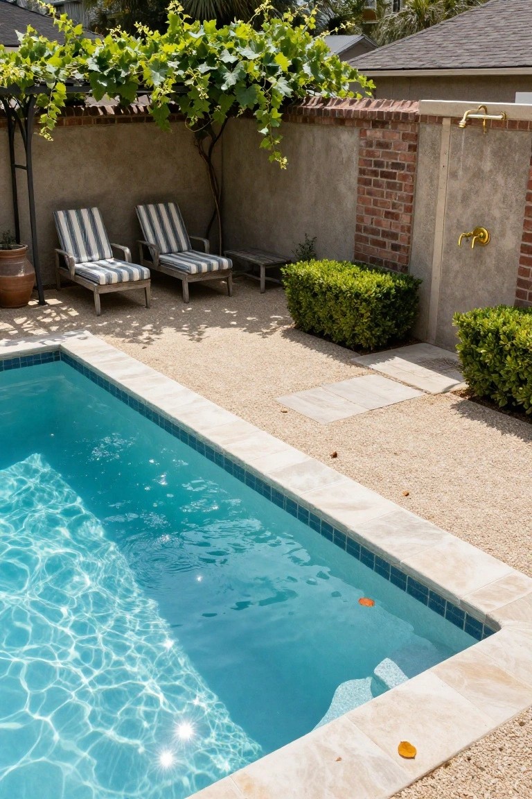 A rectangular swimming pool with turquoise water sits beside a gravel patio that holds two striped lounge chairs under a vine-covered trellis, with green shrubs, stone pavers, and a wall-mounted outdoor shower visible in the background.