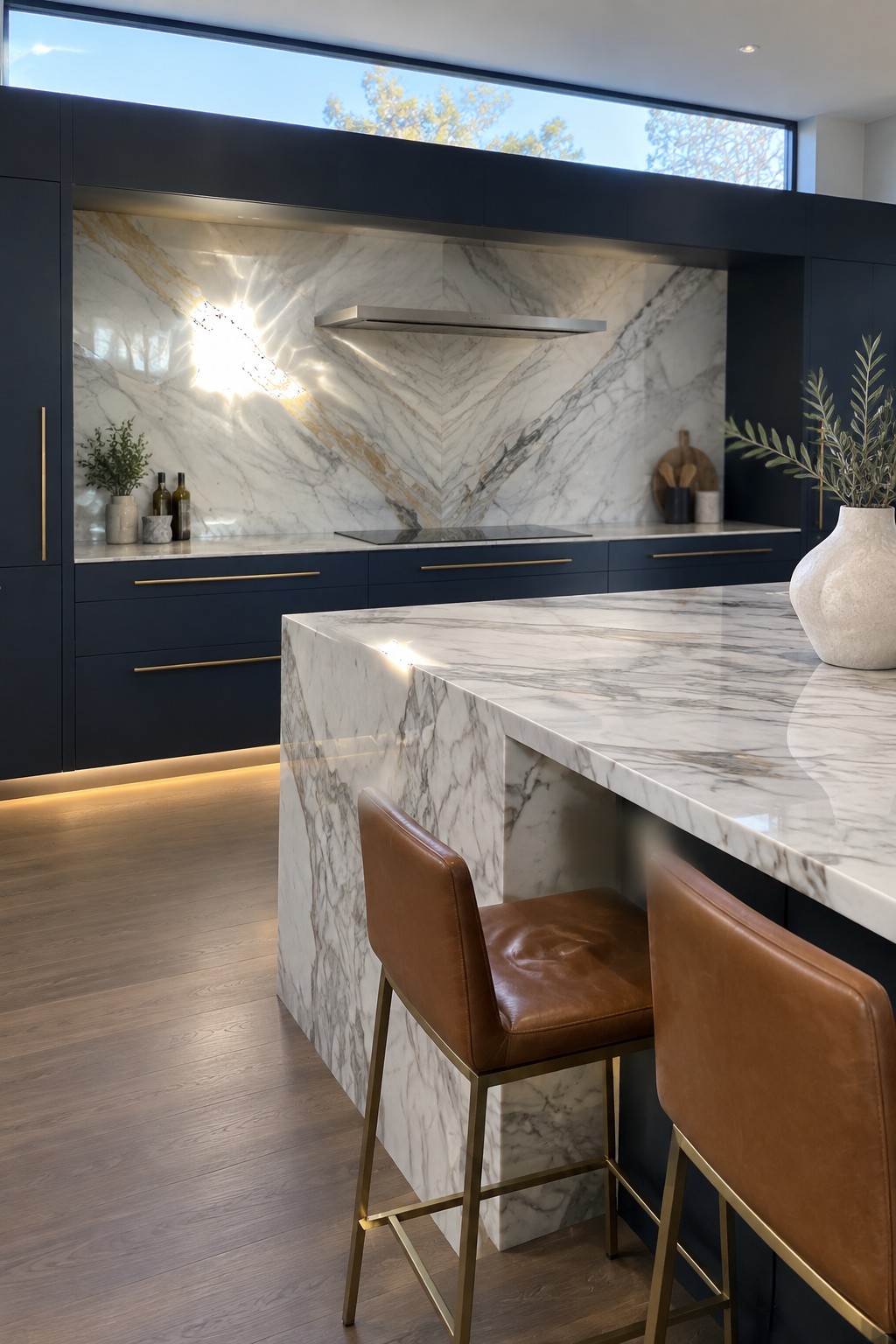 A modern kitchen with a large marble backsplash showing dramatic veining above the cooktop, dark navy cabinets with brass pulls, and a marble island with brown leather bar stools.
