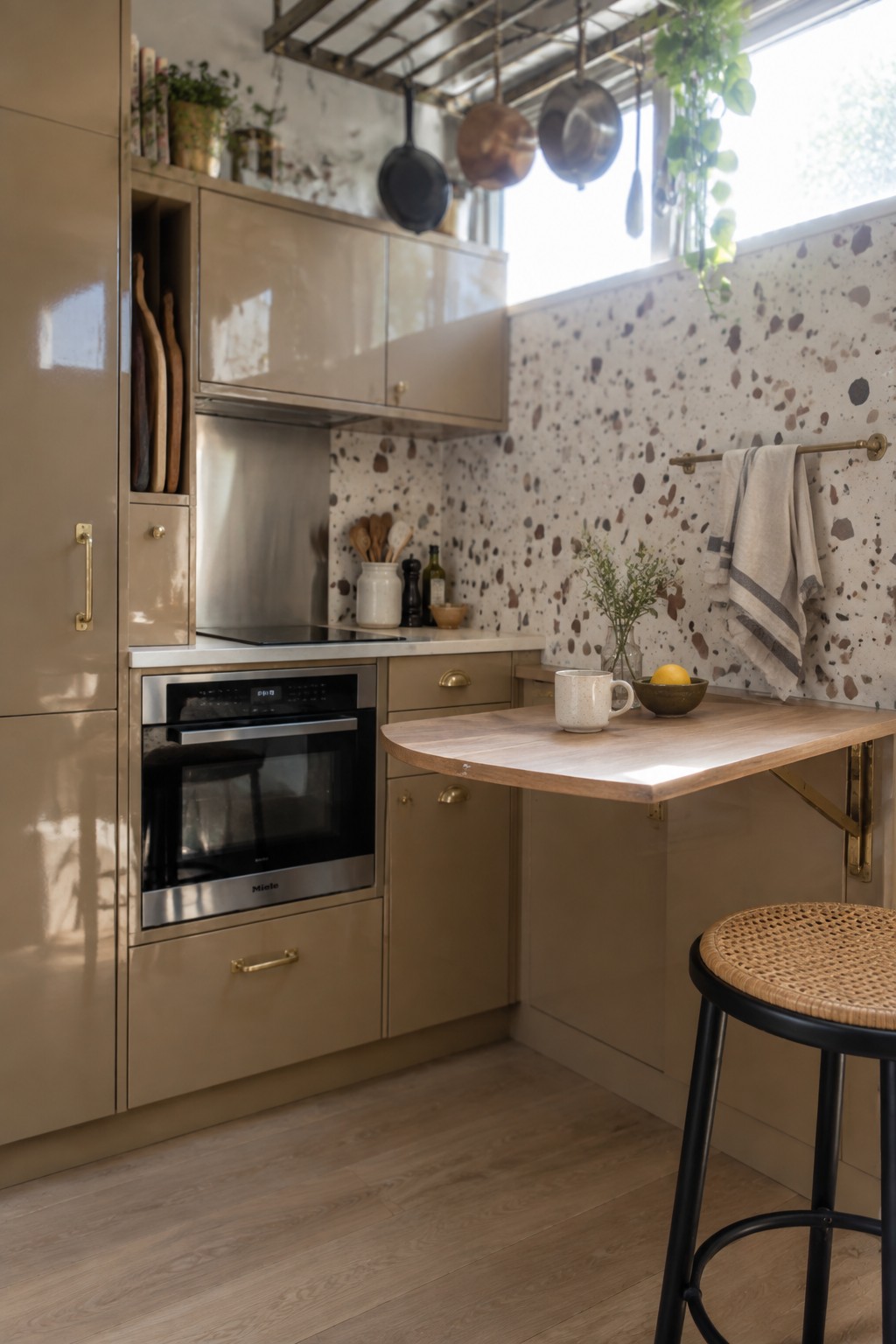 A small kitchen with beige cabinets, a built-in oven, a fold-down wooden table extension on the right, hanging pots and pans from a ceiling rack, and a speckled terrazzo wall.