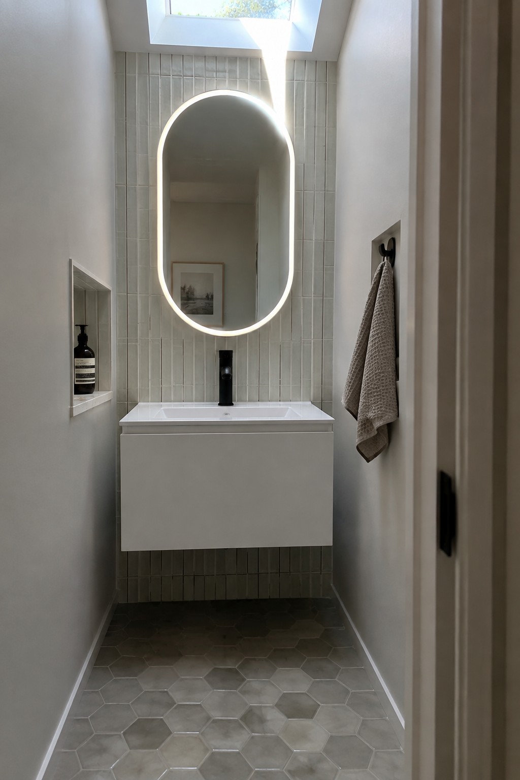 A narrow bathroom with light gray vertical tiles, a floating white vanity, a backlit oval mirror, a recessed niche holding a soap bottle, and hexagonal floor tiles.