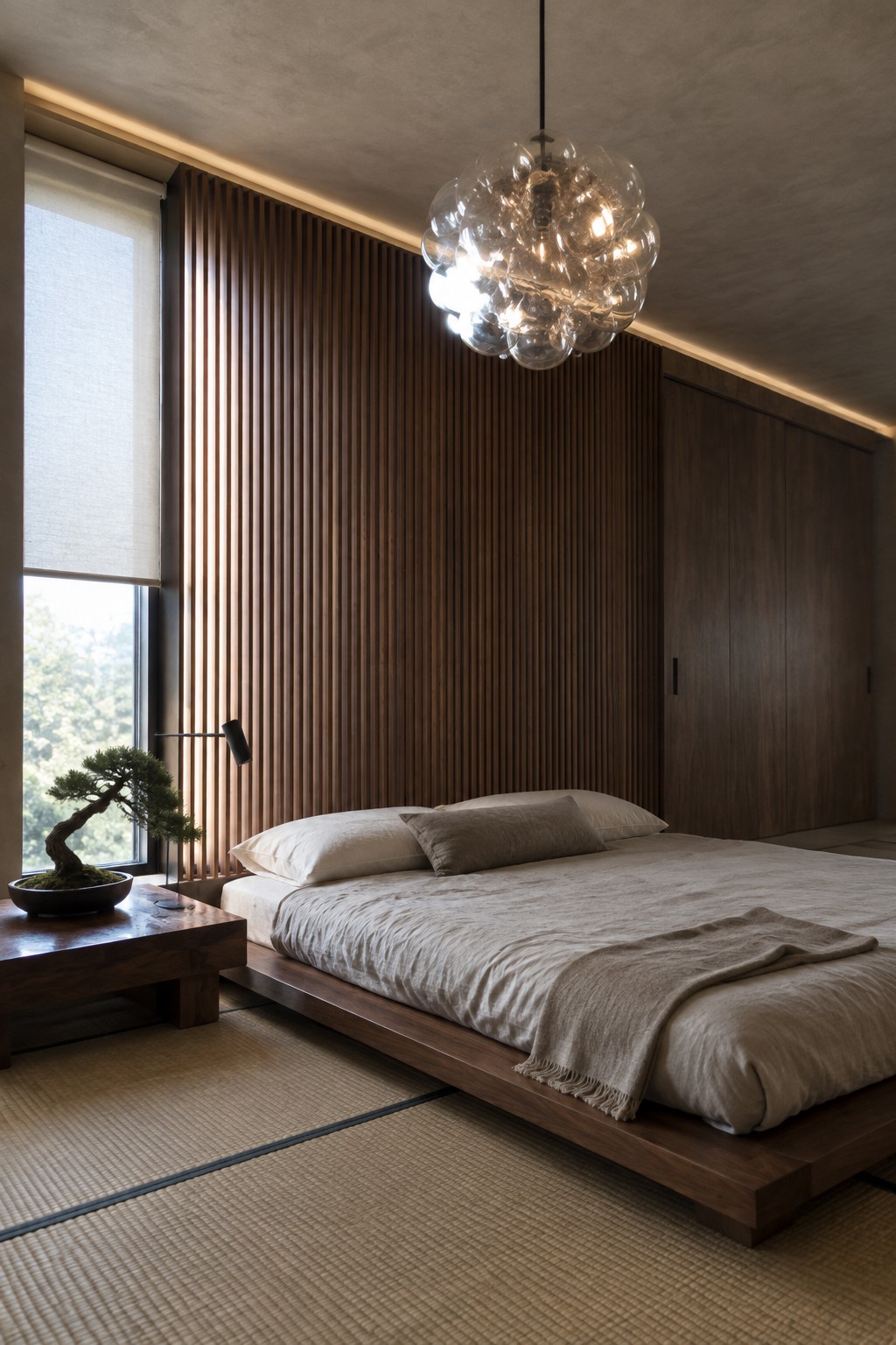 A modern bedroom with a low wooden platform bed dressed in neutral linens, a large bubble-style glass chandelier overhead, and a full wall of vertical dark wood slats beside a window with a bonsai on a side table.