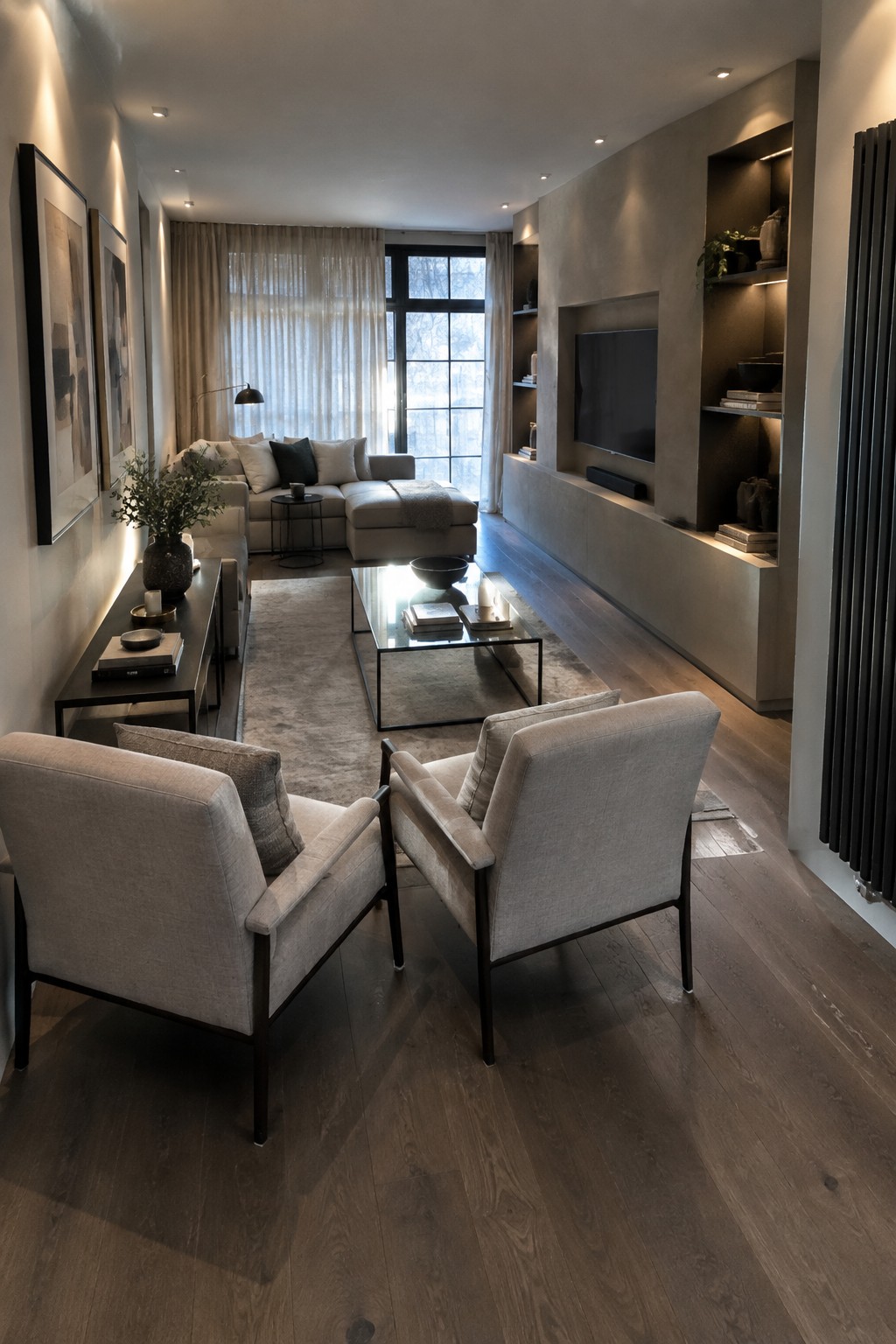 A long modern living room with two light upholstered armchairs facing a glass coffee table in the foreground and a sectional sofa positioned further back near a large window.