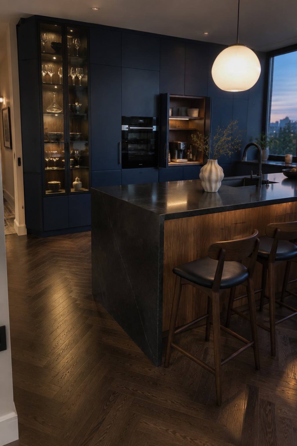 A modern kitchen featuring full-height dark navy cabinetry with glass doors, a black stone island with wooden bar stools, and warm under-cabinet lighting.