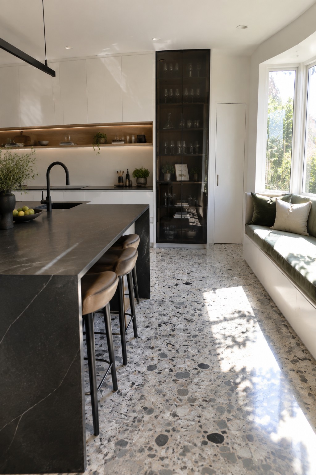 A modern kitchen interior featuring white full-height cabinets along the back wall, a tall dark glass cabinet, a dark stone island with four brown leather stools, and a built-in window seat on the right.