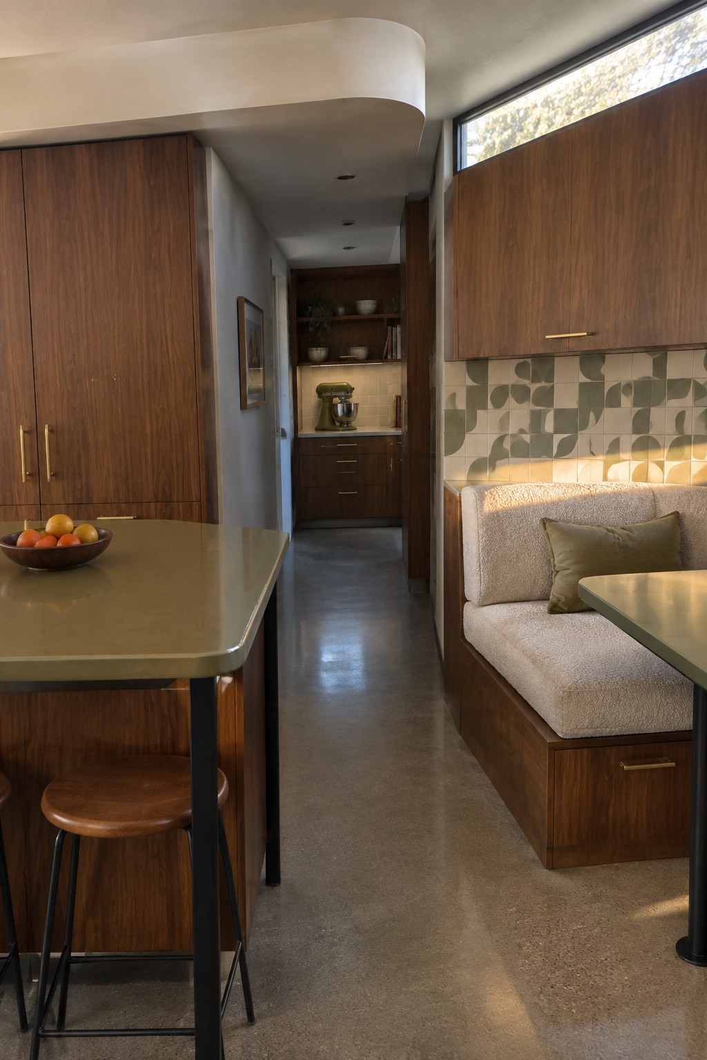 Interior view of a modern kitchen and dining area with a green island and wooden stools in the foreground, a built-in upholstered bench along the right wall, and a hallway leading to a closed kitchen with dark wood cabinets and a green stand mixer.