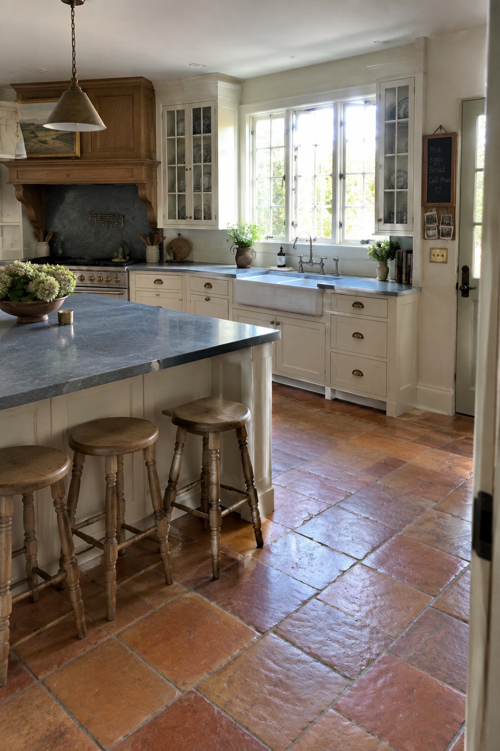A kitchen interior featuring off-white shaker cabinets, a large island with blue-gray stone countertop, three wooden stools, a farmhouse sink under windows, and terracotta tile flooring.