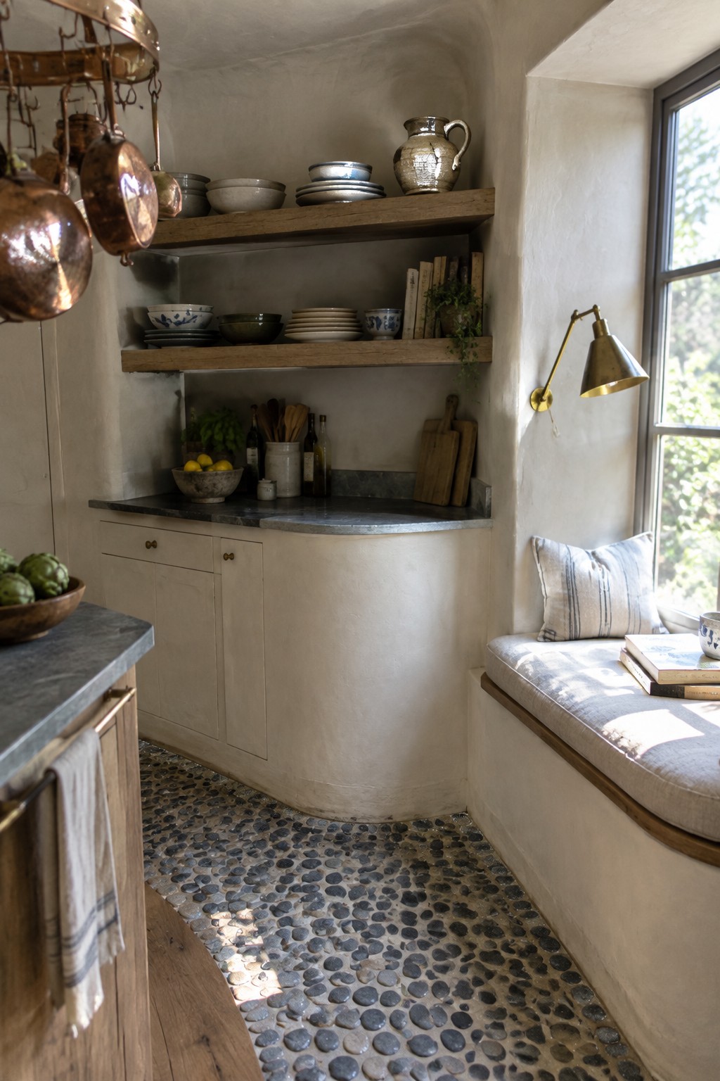 Rustic kitchen with hanging copper pots, wooden shelves, pebble floor, and window seat.