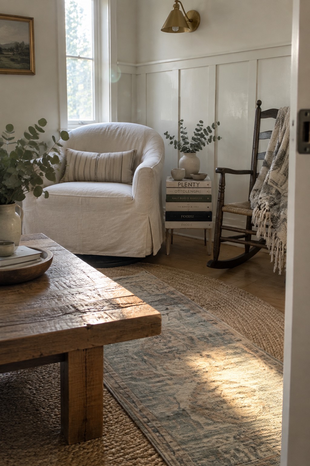 A living room with a white slipcovered armchair, a dark wooden rocking chair with a blanket, a rustic wooden coffee table, and two layered rugs on a wood floor near a window with natural light.