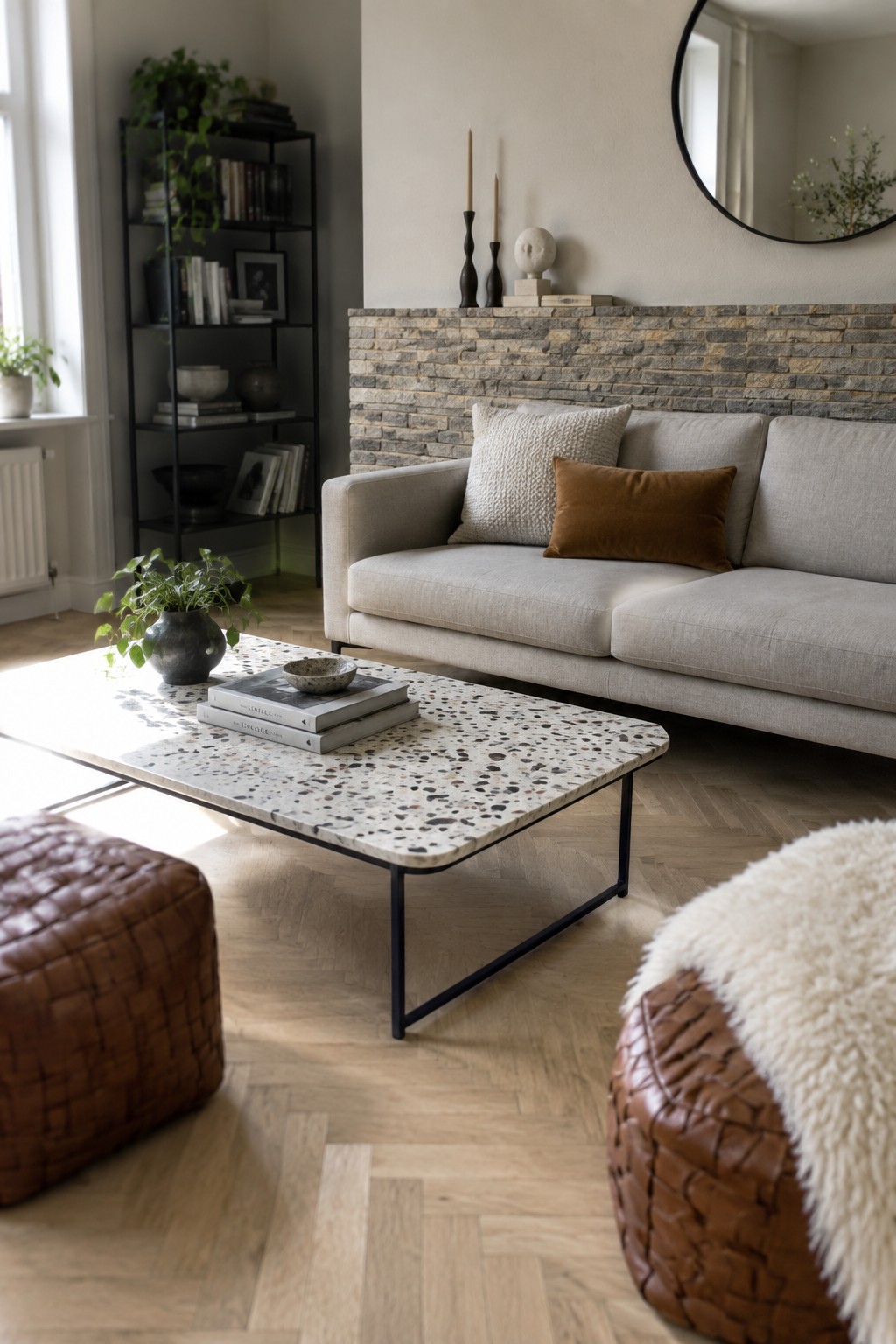 A living room with a light gray sofa in front of a stacked stone accent wall, a terrazzo coffee table, and two brown leather poufs on a herringbone wood floor.