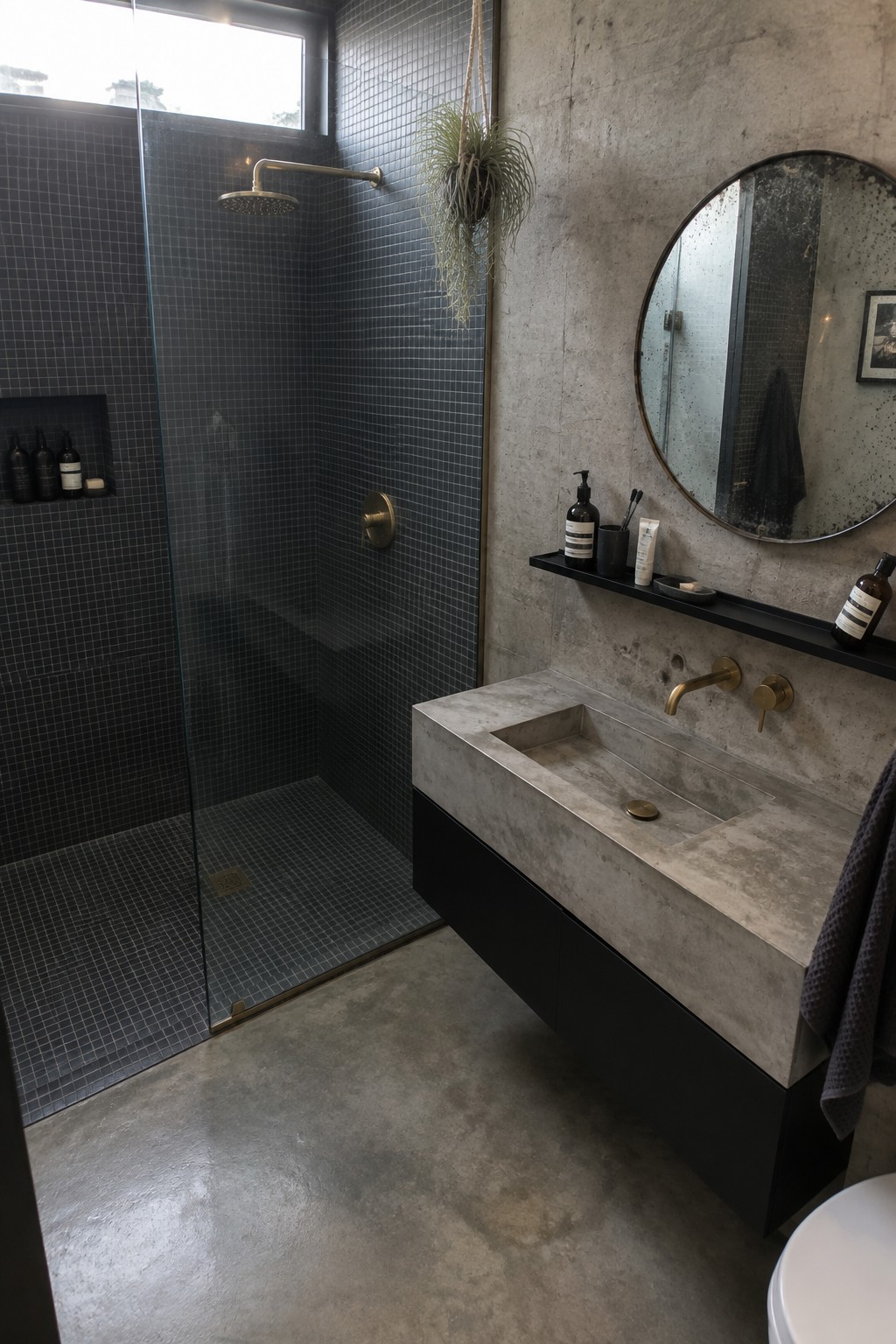 A modern bathroom featuring a dark mosaic tiled walk-in shower beside a floating concrete vanity with brass fixtures.