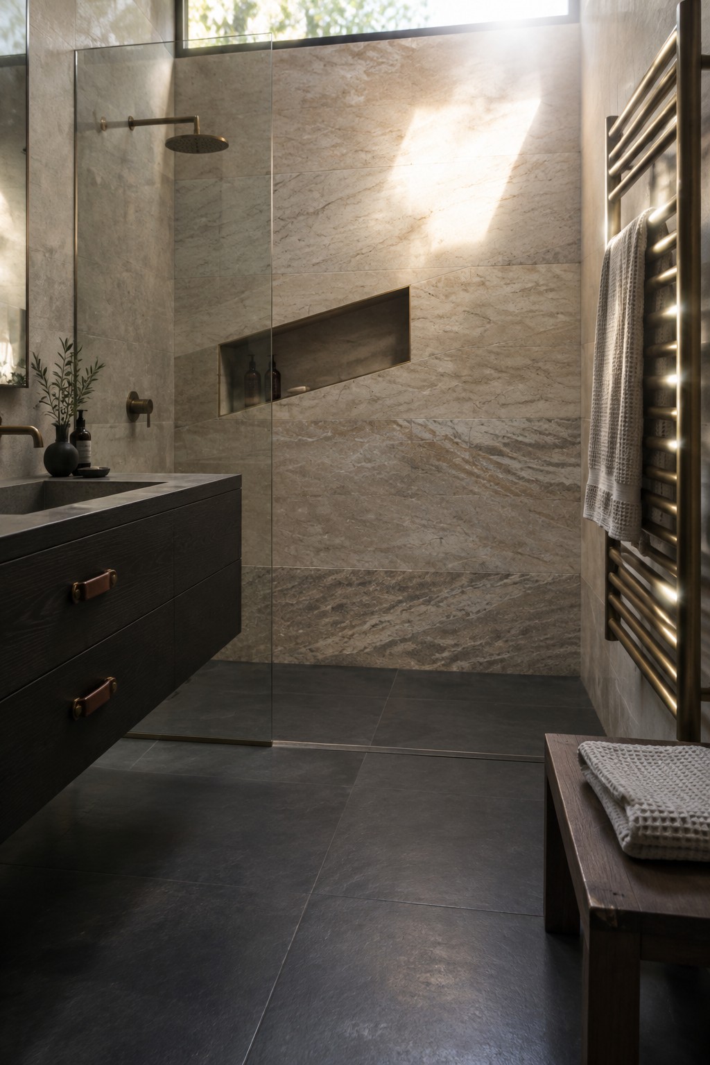 A modern bathroom with a glass-enclosed walk-in shower featuring large beige marble tiles, a recessed niche holding bottles, dark floor tiles, a floating dark wood vanity, and a brass towel warmer on the right wall.