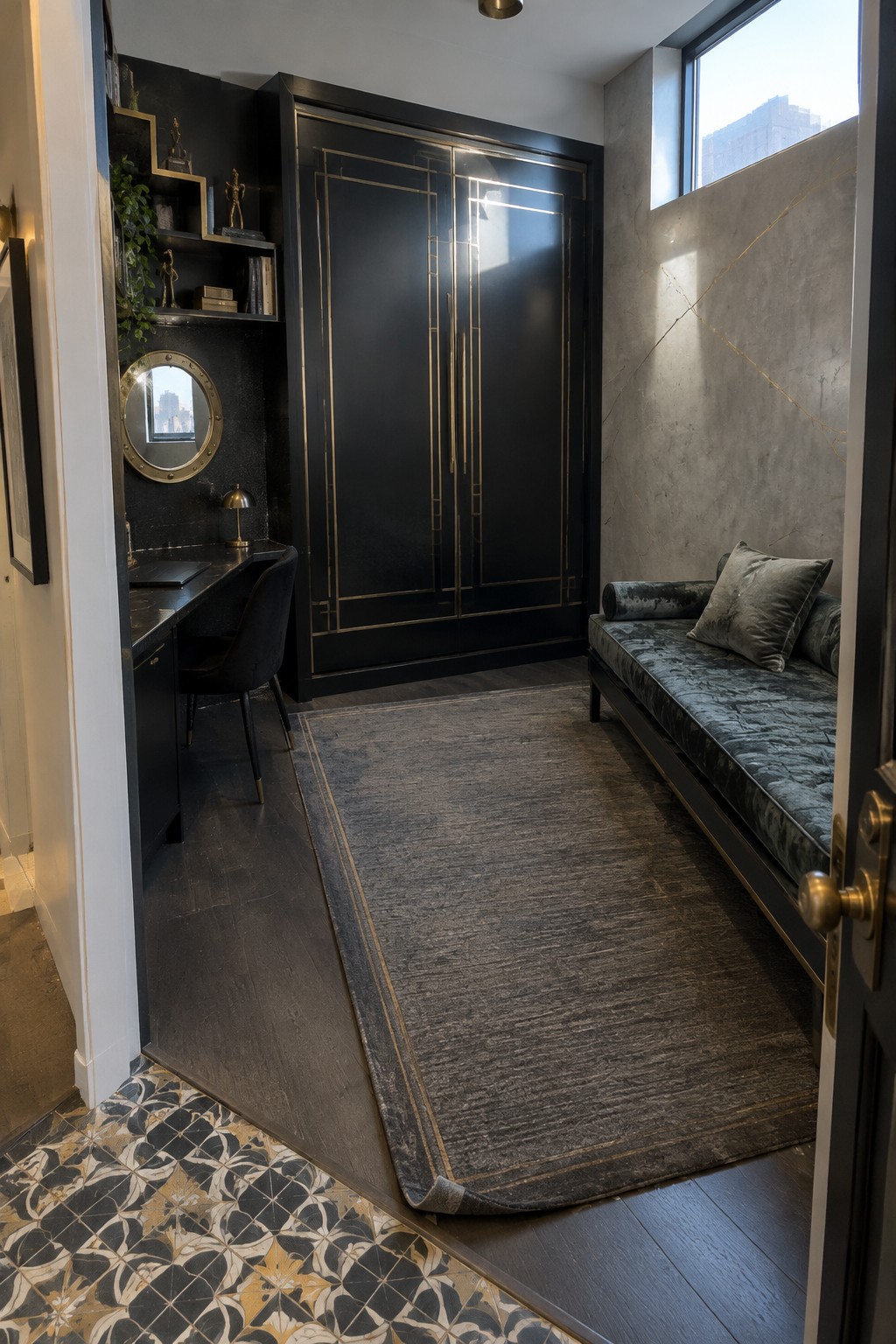 An interior view of a bedroom with a large black wardrobe trimmed in gold, a gray velvet daybed, a small desk area, and a large area rug on dark wood flooring.