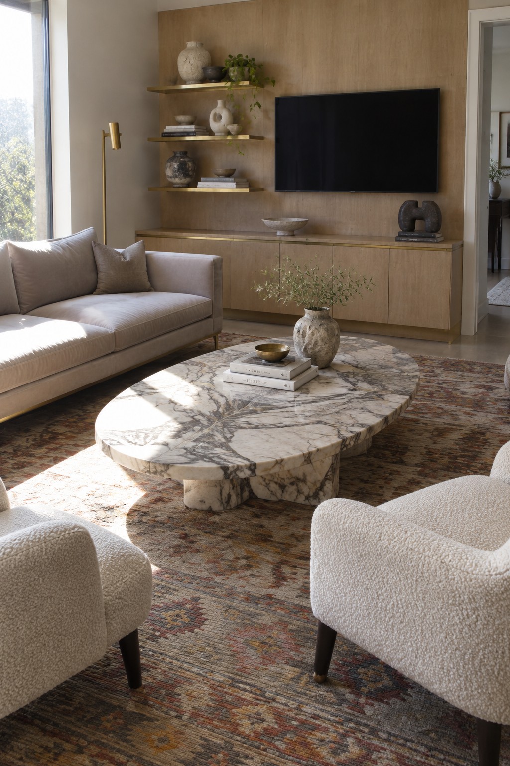 A living room with a large oval marble coffee table centered on a patterned rug, surrounded by a light gray sofa, two white boucle armchairs, a brass-trimmed wood console, and floating shelves with decor.