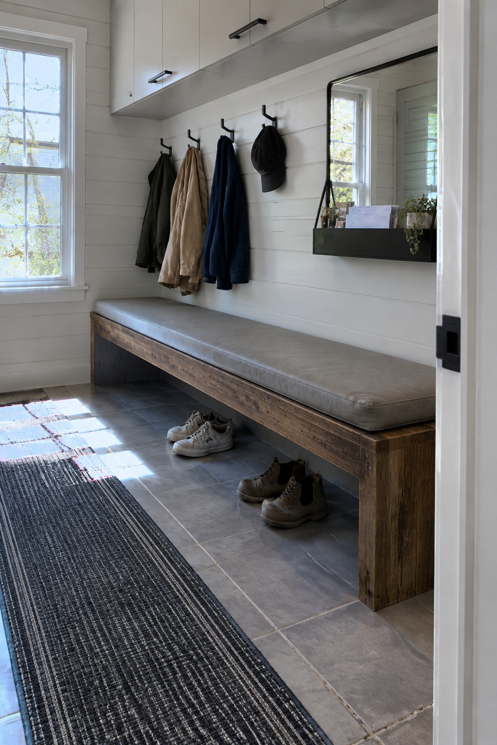 A long wooden bench with a gray cushion is mounted below black hooks holding coats and a cap in a white shiplap entryway with a mirror above a black shelf.