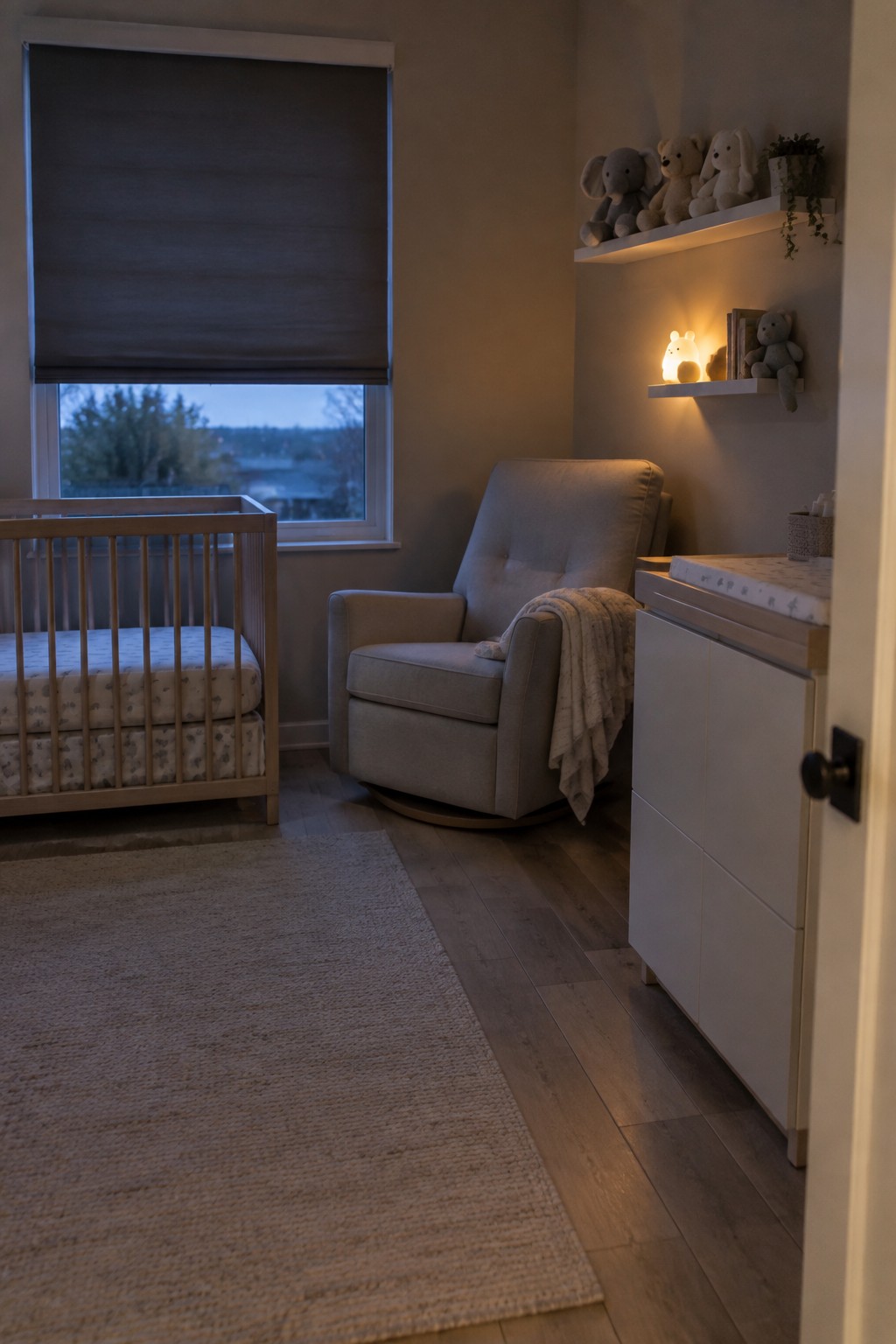 A nursery interior with a wooden crib, gray upholstered rocking chair, white dresser, woven rug on wood flooring, and floating shelves holding stuffed animals and a night light.