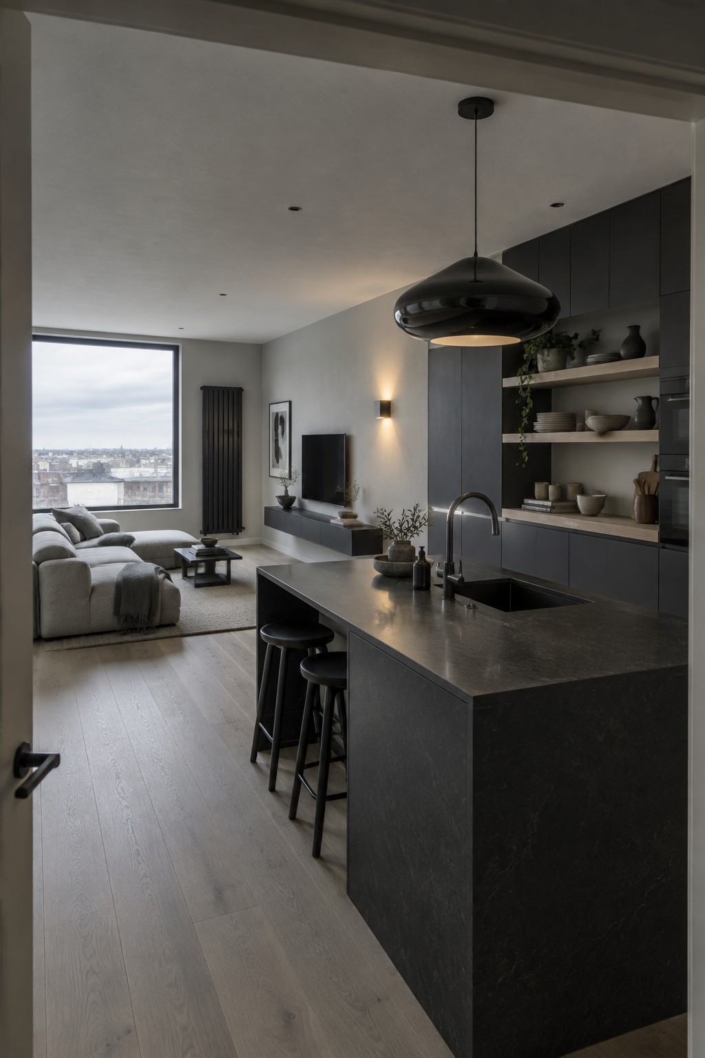 A modern apartment interior showing a dark stone kitchen island with black cabinetry, open shelving, and a living area with sofa and large window beyond.