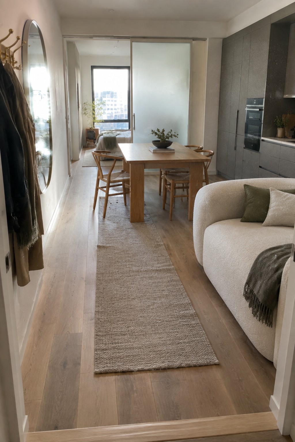 An apartment interior with a wooden dining table on a long runner rug, a curved sofa to the right, gray kitchen cabinets, and frosted sliding doors partially open to reveal a bedroom.