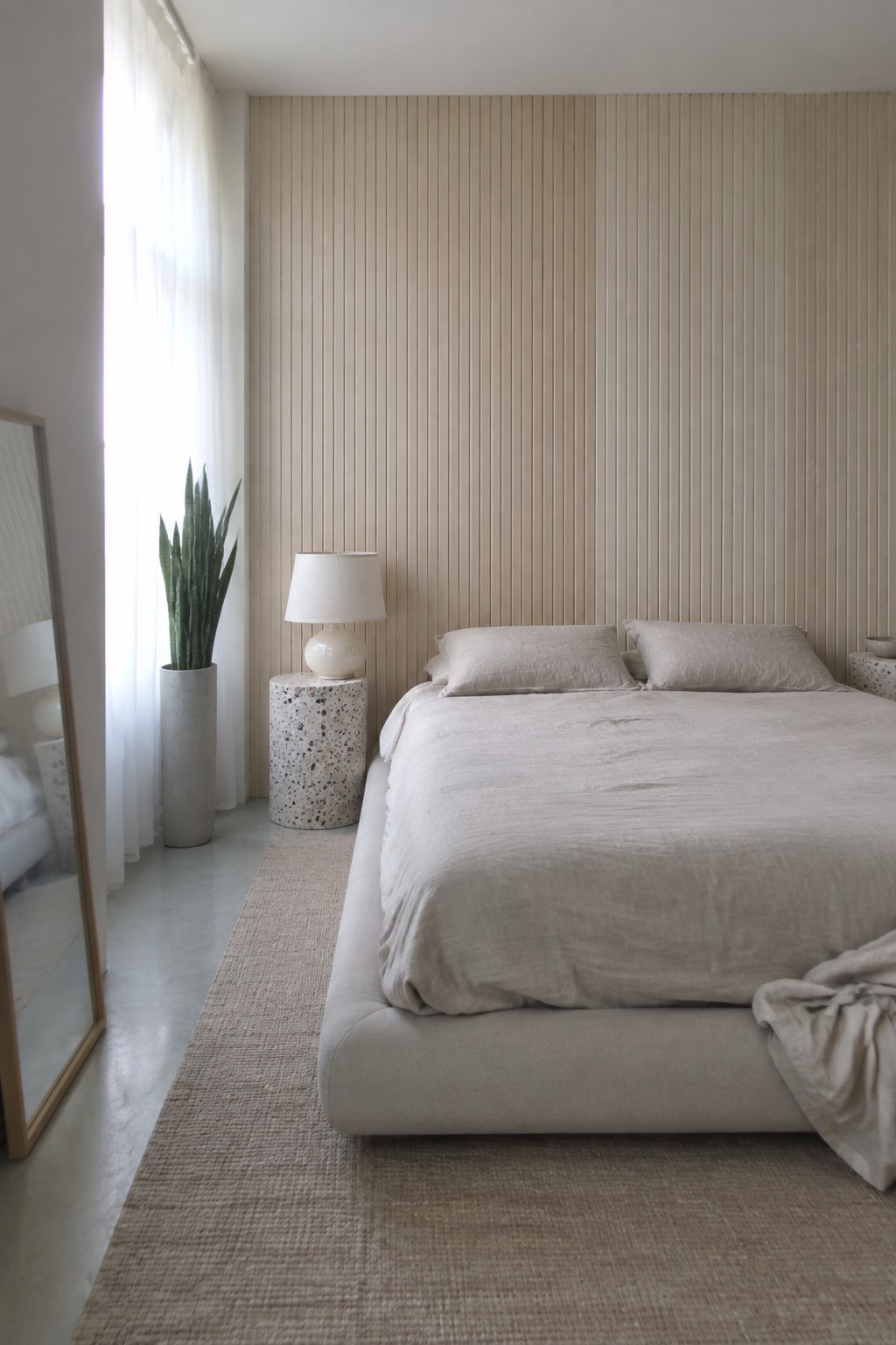A bedroom with a large bed dressed in light linen, positioned against a wall covered in vertical light wood fluted panels, with a terrazzo side table and potted plant to the left.