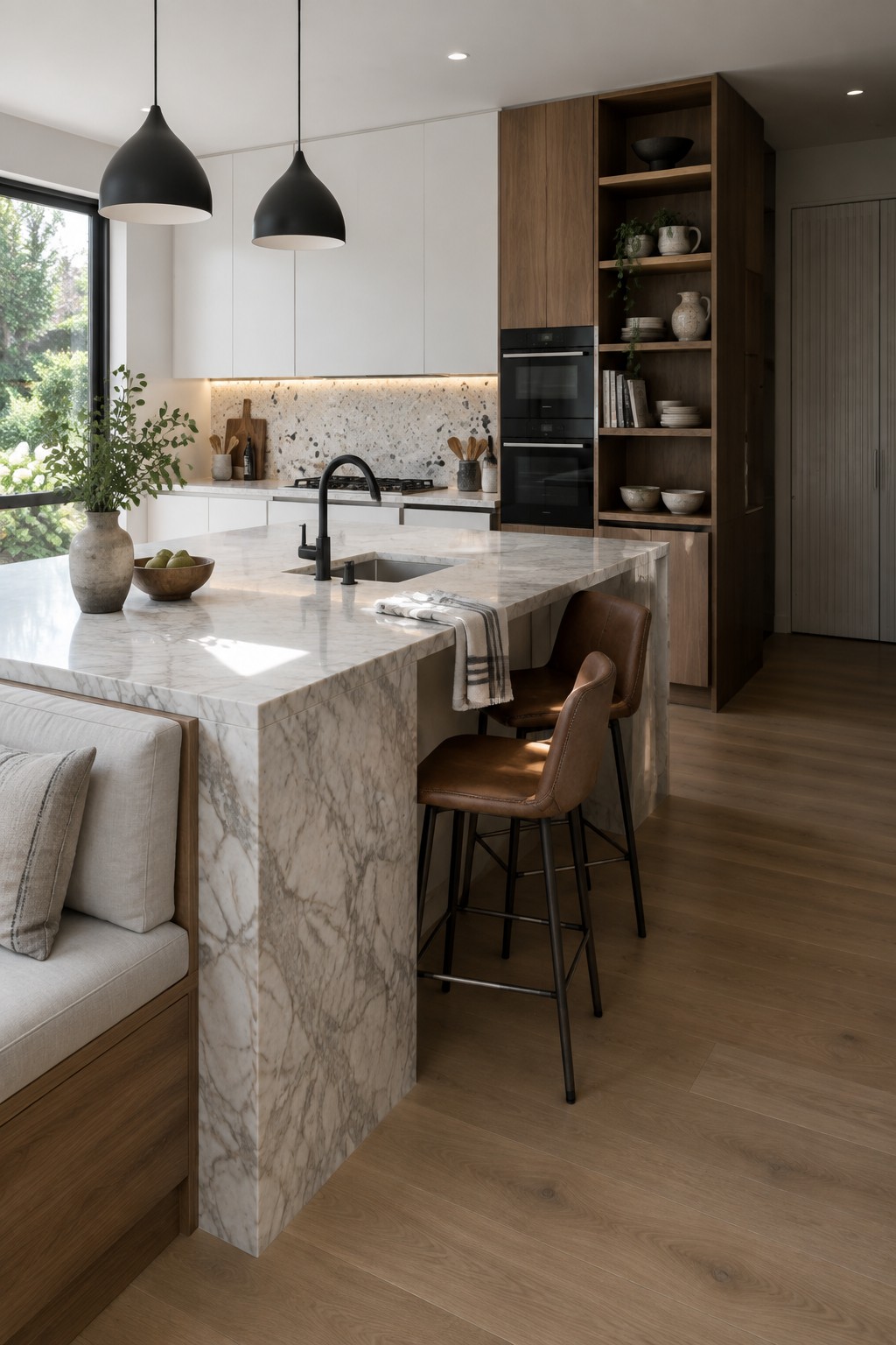 A modern kitchen with a large marble island featuring a built-in upholstered bench on one side and two brown leather bar stools on the other, surrounded by white cabinetry and dark wood open shelving.