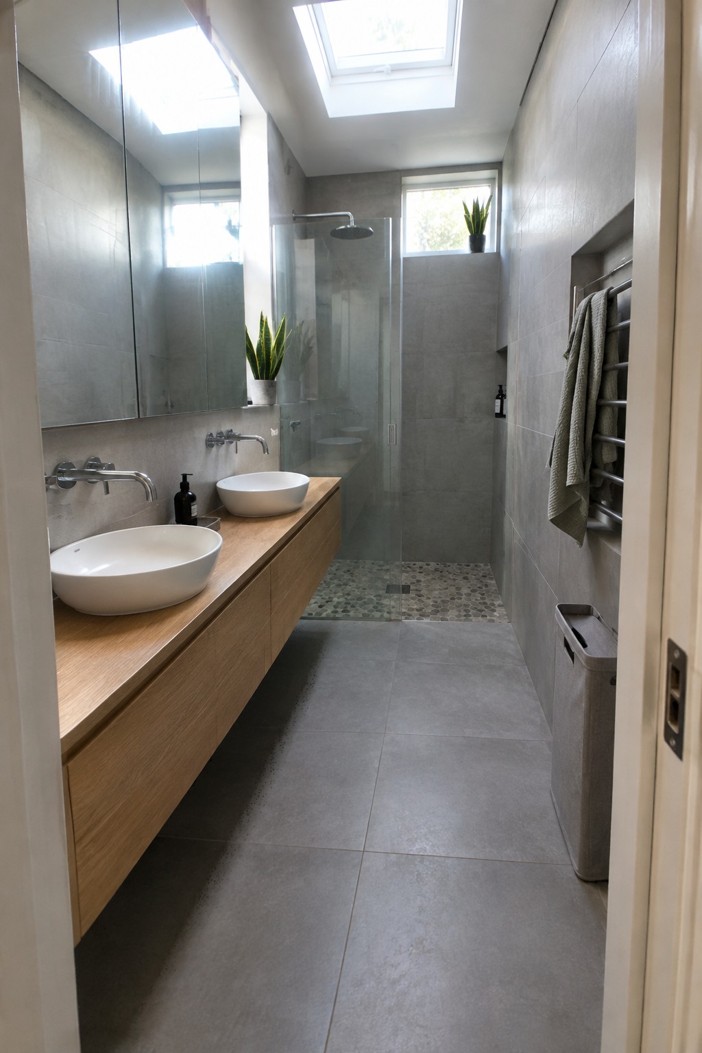 A modern bathroom with a long floating wooden vanity holding two white vessel sinks beneath a large mirror, gray tiled walls, and a glass-enclosed shower at the end of the narrow room.