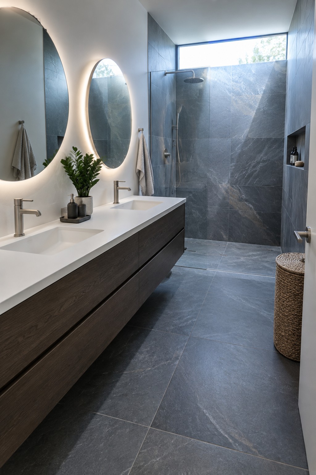 A modern bathroom with a long floating double vanity in dark wood, white countertop, two round backlit mirrors, and a gray tiled shower area.