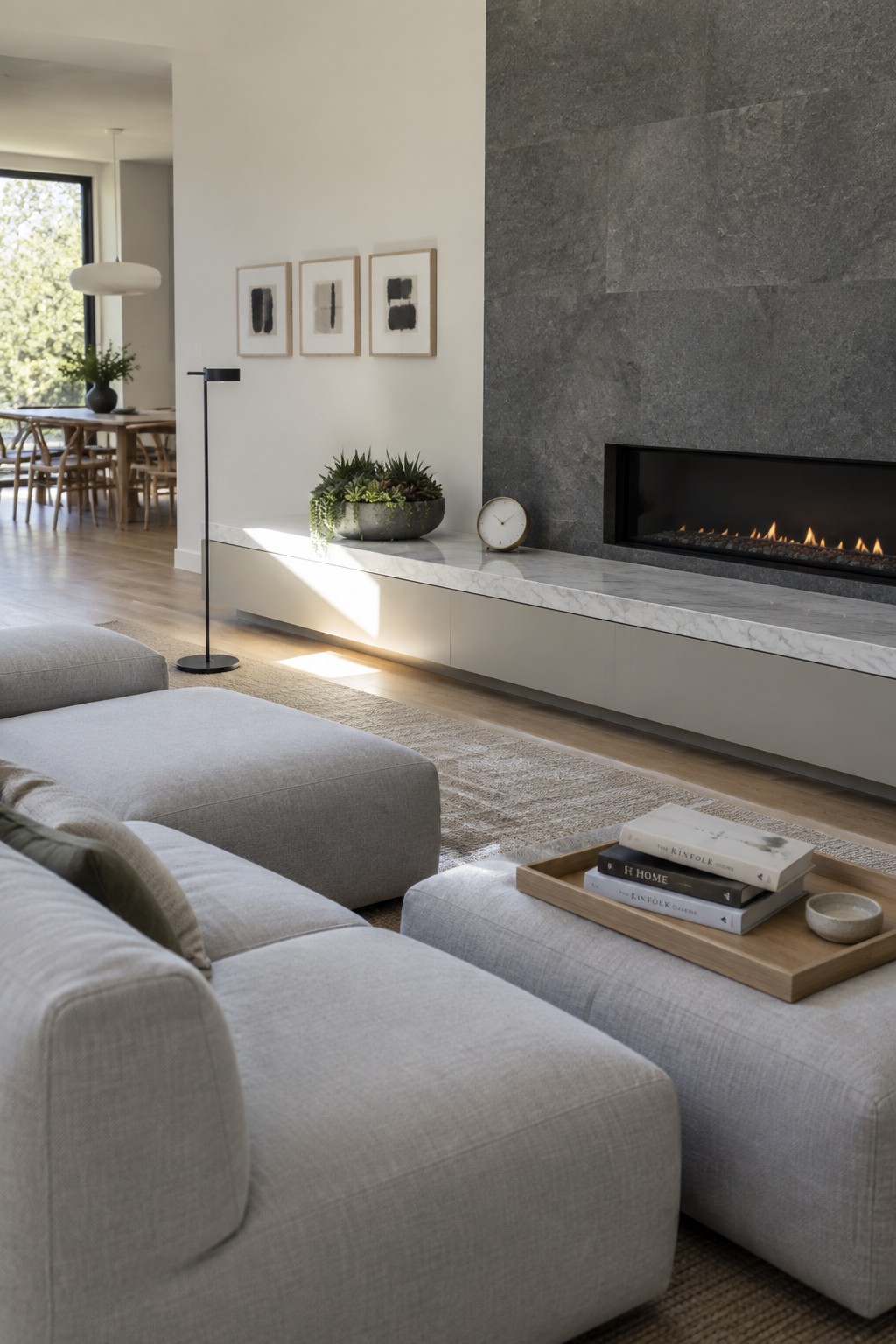 A minimalist living room with a low-profile gray sectional sofa, a floating marble console under a stone fireplace, and a wooden tray holding books on an ottoman.