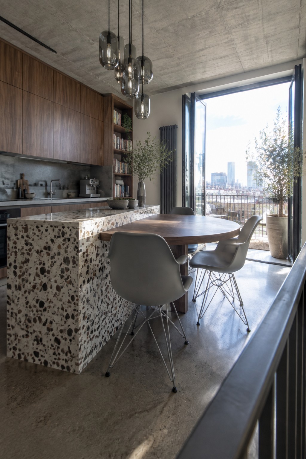 A modern open kitchen with a speckled terrazzo peninsula next to a round wooden dining table surrounded by gray molded chairs, with large glass doors opening to a balcony.