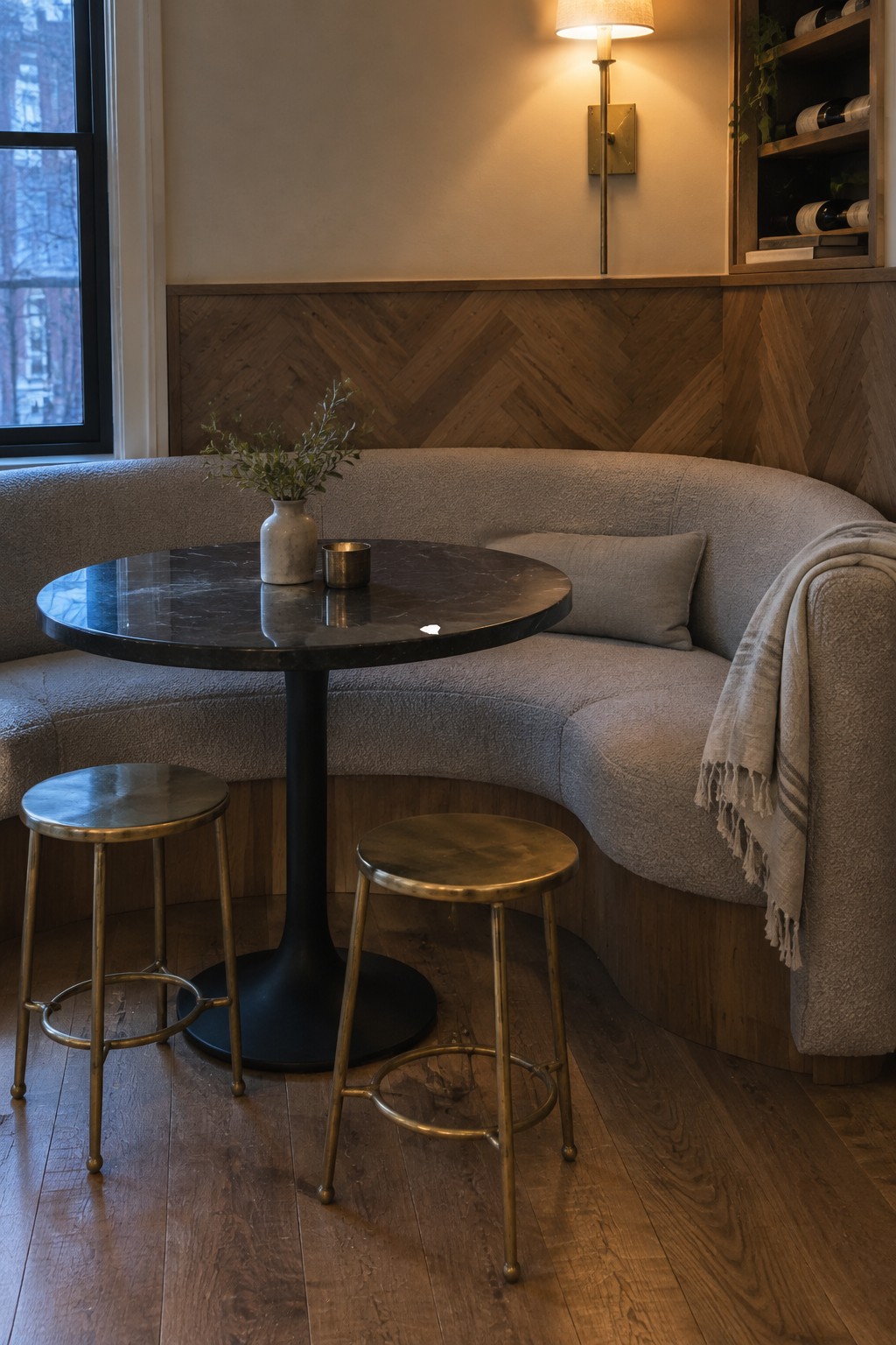 A round dark marble dining table with two brass stools sits in front of a curved light gray upholstered banquette against wood herringbone paneling.
