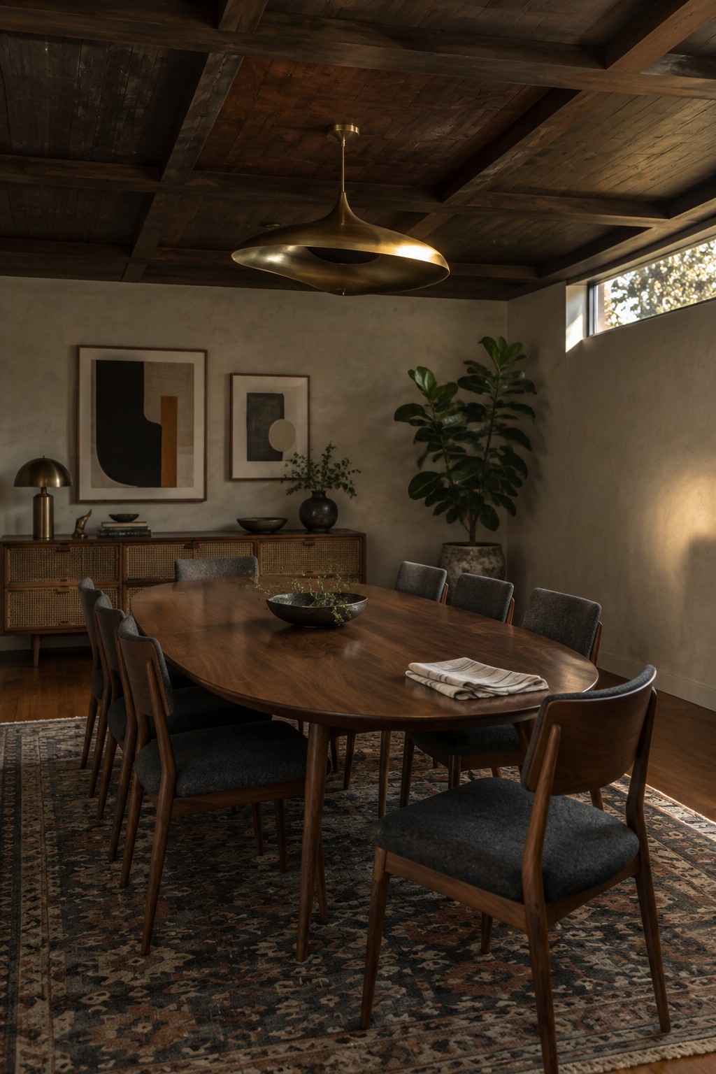 A dining room featuring a long wooden table with upholstered chairs on a patterned rug beneath a dark wood ceiling with exposed beams and a large brass pendant light.