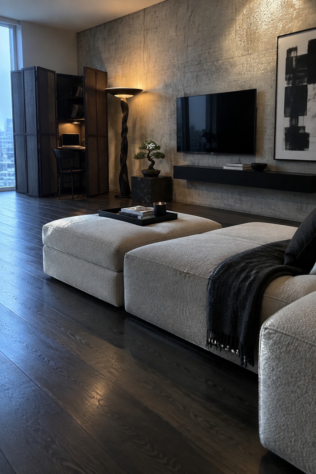 A modern living room with two large beige modular ottomans on dark wood flooring, a wall-mounted TV on a concrete wall, a twisted floor lamp, and a small desk area in the background.