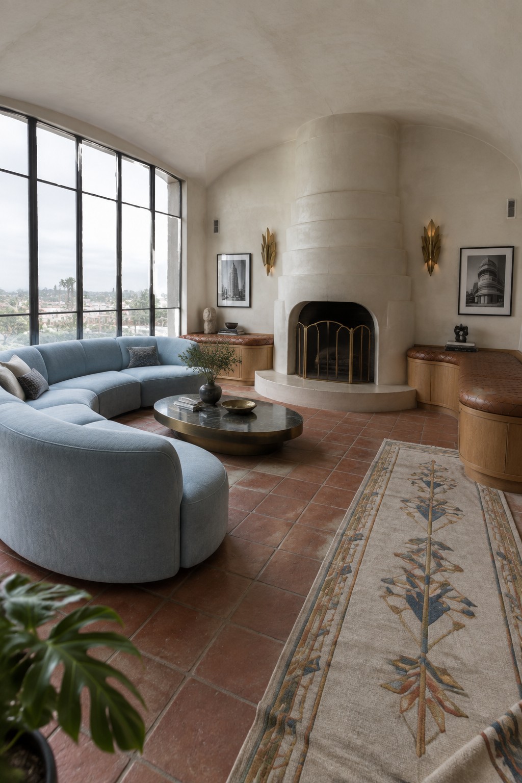 A living room featuring a large curved blue sectional sofa, a round dark coffee table, a curved plaster fireplace with brass screen, and tall black-framed windows on the left.