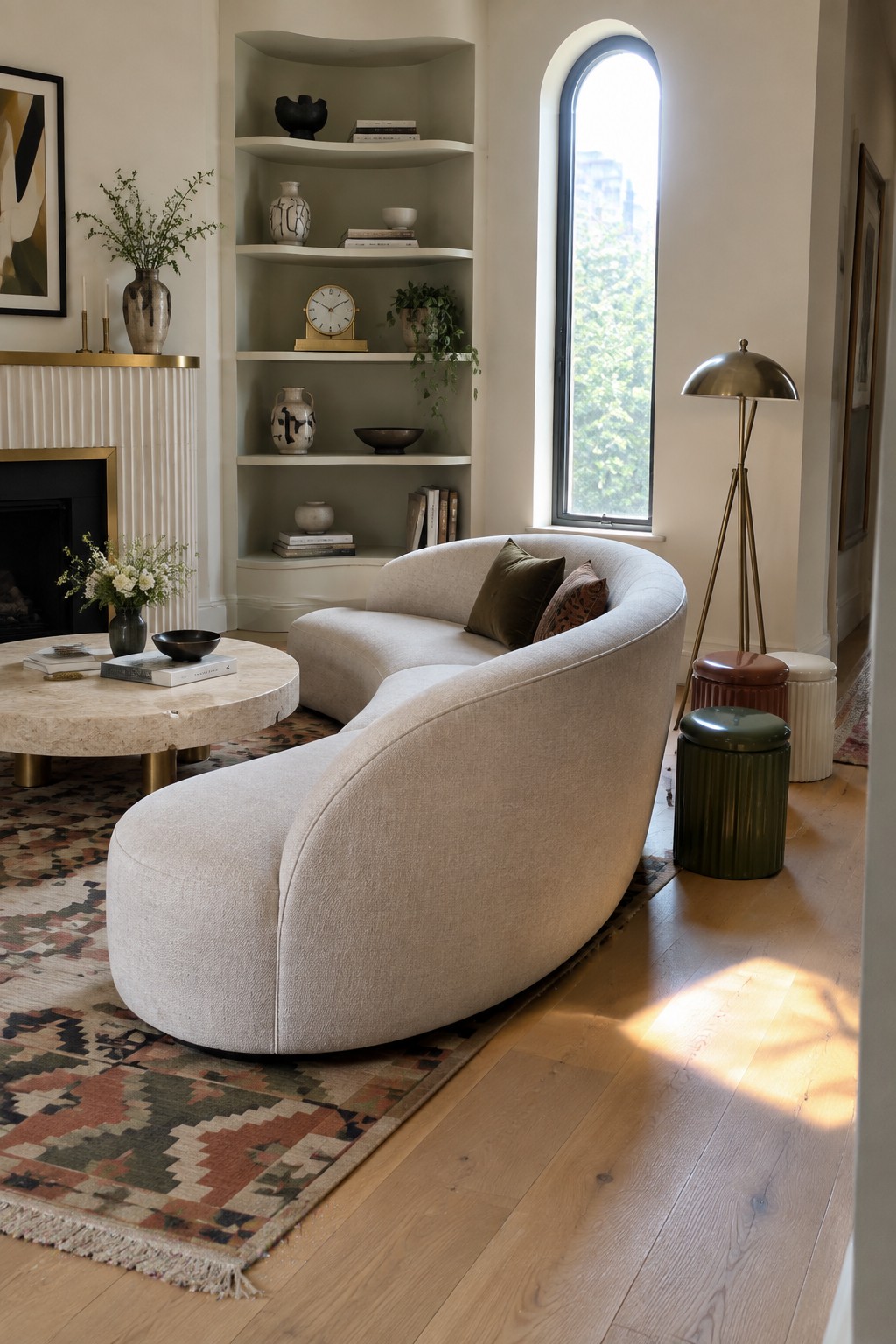 A living room with a large curved beige sofa on a patterned rug, built-in shelves with decor, a round marble coffee table, and a brass tripod floor lamp near an arched window.