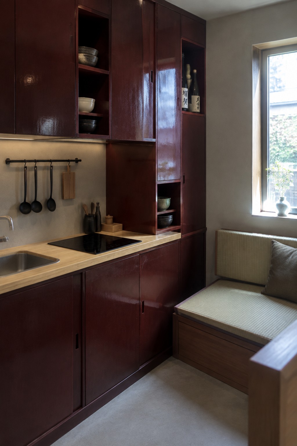 A compact kitchen with tall dark red cabinets, light wood countertops, a black induction cooktop, and a built-in bench beside a window.