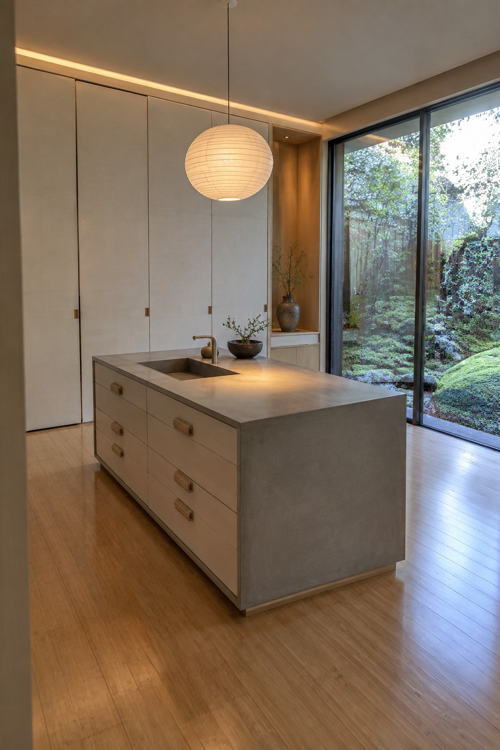 A modern kitchen with a large concrete island, light wood drawers with woven handles, a sink, and full height cabinets on the left wall beside tall glass doors to a garden.